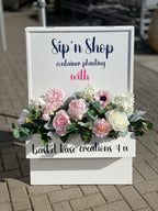 Decorative white wooden sign floral box, accented with white roses, pink peonies, blush anenome, pink dahlias, white hydrangeas, dusty miller leaves and eucalyptus varieties floral arrangement displayed at the entrance to a demonstration event.