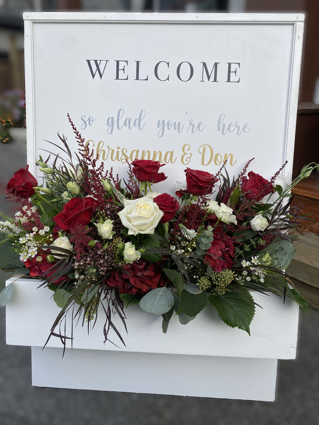 Decorative white wooden 'WELCOME' sign floral box, accented with red and white rose, hydrangea, astilbe, lisianthus, wax flower and eucalyptus varieties floral arrangement displayed at the entrance to a wedding ceremony.