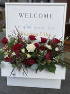 Decorative white wooden 'WELCOME' sign floral box, accented with red and white rose, hydrangea, astilbe, lisianthus, wax flower and eucalyptus varieties floral arrangement displayed at the entrance to a wedding ceremony.
