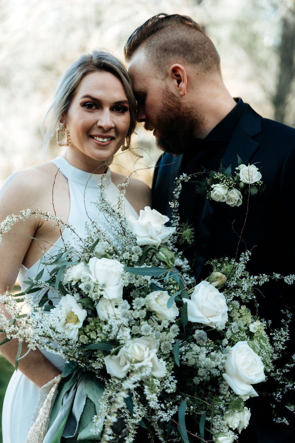 A bride and groom standing together. The bride holds a large, textured white and green bouquet featuring garden roses, poppies, spirea, tulips, and astrantia. Long streamers of taupe satin, green hand-dyed silk, and champagne gold sequins trail from the stems.
