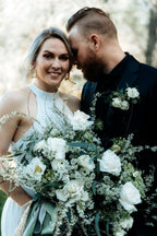 A bride and groom standing together. The bride holds a large, textured white and green bouquet featuring garden roses, poppies, spirea, tulips, and astrantia. Long streamers of taupe satin, green hand-dyed silk, and champagne gold sequins trail from the stems.