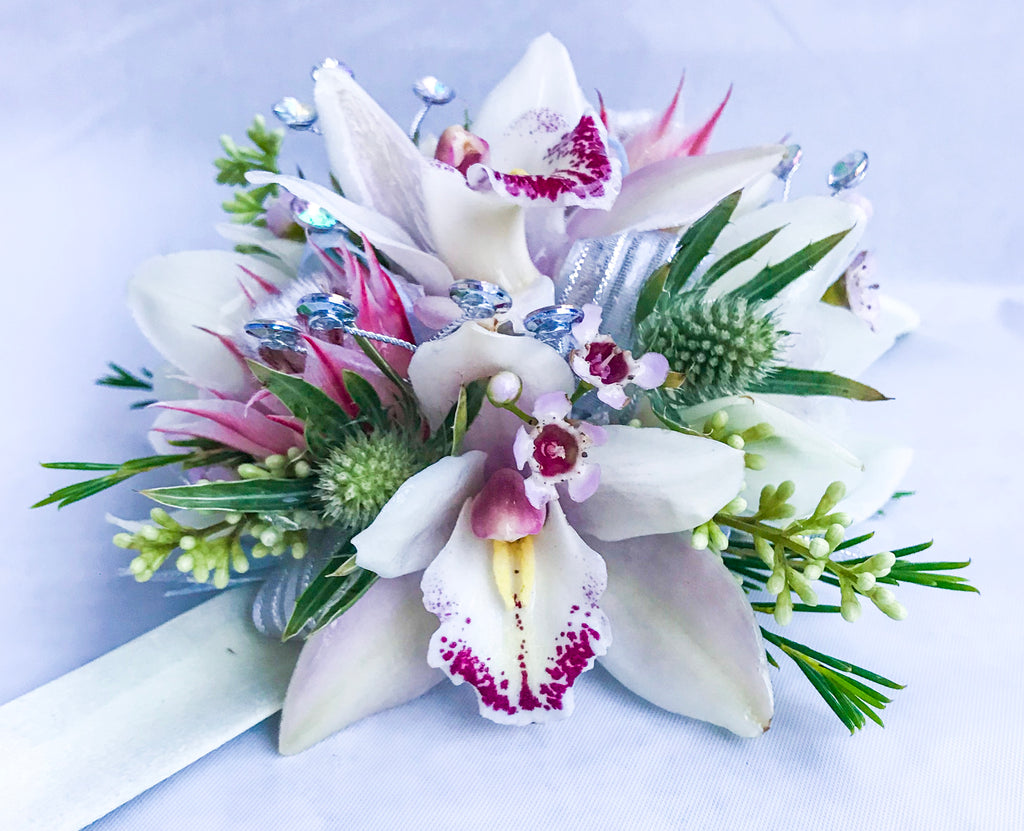 A floral wrist corsage on a white slap band featuring white cymbidium orchids. Accents include pink blushing bride protea, green eryngium, seeded eucalyptus, finished with silver striped and iridescent organza ribbon loops and shimmering silver rhinestone sprays