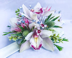 A floral wrist corsage on a white slap band featuring white cymbidium orchids. Accents include pink blushing bride protea, green eryngium, seeded eucalyptus, finished with silver striped and iridescent organza ribbon loops and shimmering silver rhinestone sprays