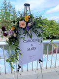 Wedding welcome sign on easel featuring a left top corner floral swag with vibrant yellow spray roses, peach garden roses, purple lisianthus, burgundy ranunculus, and cascading smilax and eucalyptus greenery. 