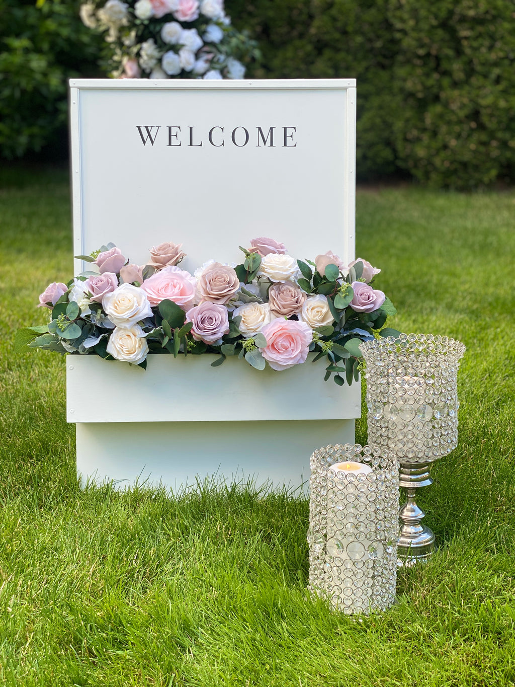 Decorative setup with 'WELCOME' sign, blush and cream rose floral arrangement, and crystal candle holders on grass.