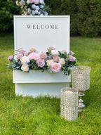 Decorative setup with 'WELCOME' sign, blush and cream rose floral arrangement, and crystal candle holders on grass.