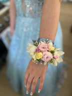 A floral wristlet corsage featuring large blush-pink ranunculus centered among white cymbidium orchids and delicate cream spray rose buds. Accented with green eryngium and scabiosa buds, dried bunny tails, seeded eucalyptus sprigs, and shimmering rhinestone sprays, all tied with a pale blue-colored satin ribbon.