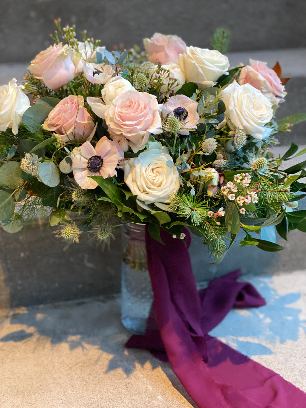 A large, lush bridal bouquet featuring white and blush garden roses, peony buds, anenome, delicate astrantia and wax flower, green-tinted eryngium, accented with douglas fir, seeded, and silver-dollar eucalyptus and mixed greenery, finished with burgundy wine coloured hand dyed silk ribbon streamers.