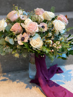 A large, lush bridal bouquet featuring white and blush garden roses, peony buds, anenome, delicate astrantia and wax flower, green-tinted eryngium, accented with douglas fir, seeded, and silver-dollar eucalyptus and mixed greenery, finished with burgundy wine coloured hand dyed silk ribbon streamers.