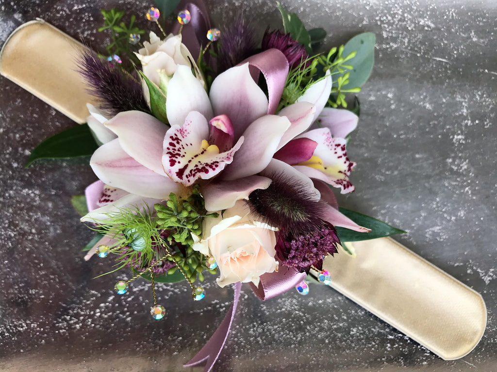 A floral wrist corsage on a gold slap band featuring a large white cymbidium orchid centered among blush spray roses and tiny red astrantia. Accents include burgundy bunny tails, nigella tips, and seeded eucalyptus, finished with mauve ribbon loops and shimmering rhinestone sprays.